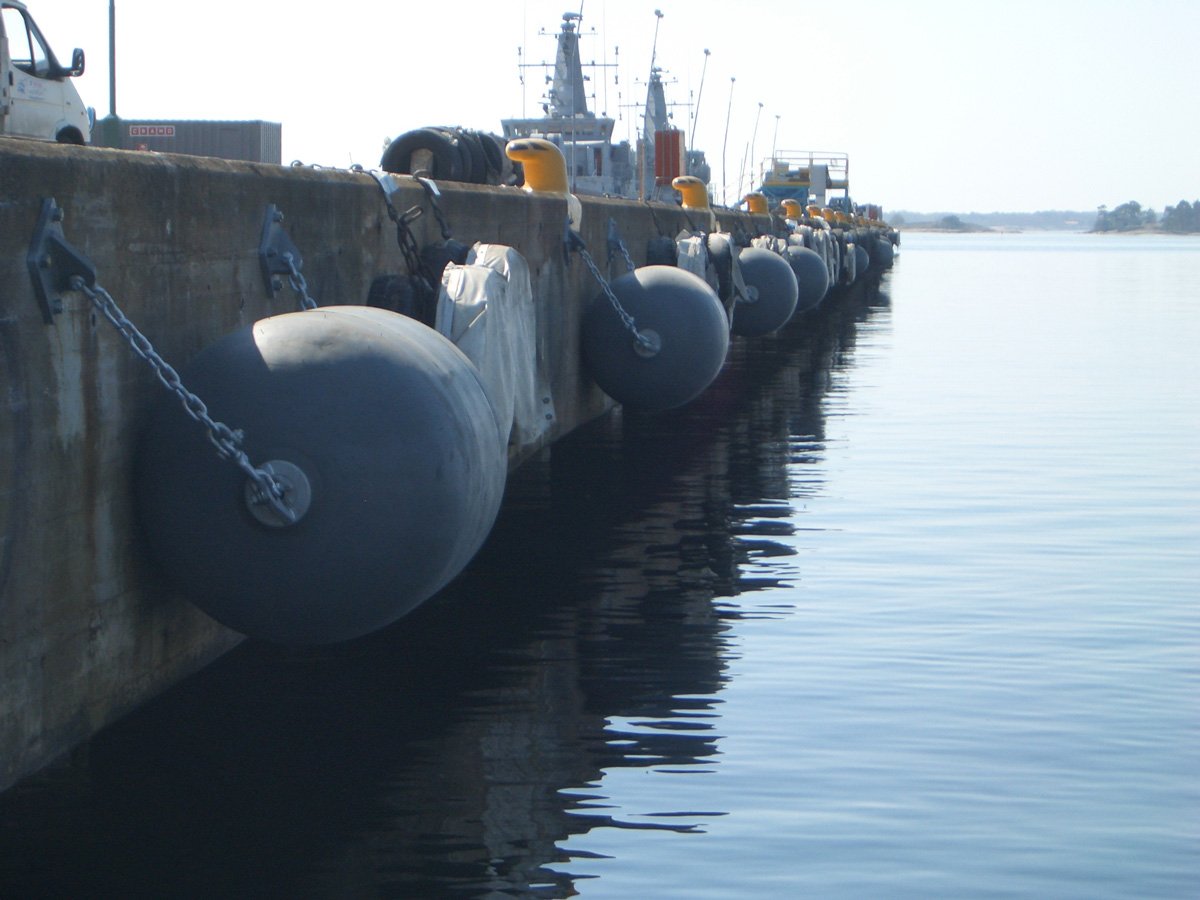 Black cylindrical foam-filled fenders secured with chain and tyre netting along a quay wall.